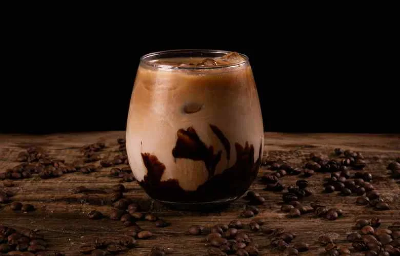 A glass of iced coffee with chocolate syrup swirls, surrounded by coffee beans on a textured wooden surface, Coffee Milkshake.