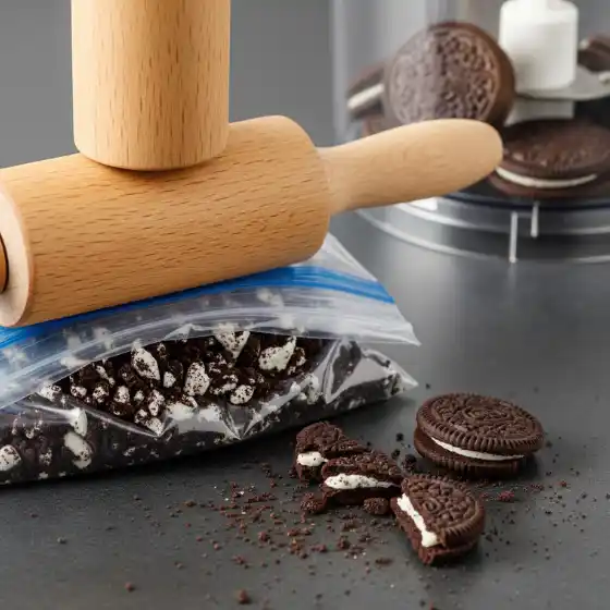 A wooden rolling pin crushes chocolate sandwich cookies inside a clear plastic bag on a dark surface, while additional whole and broken cookies lie nearby—perfect for a treat break after exploring trending Edgar haircuts for men.