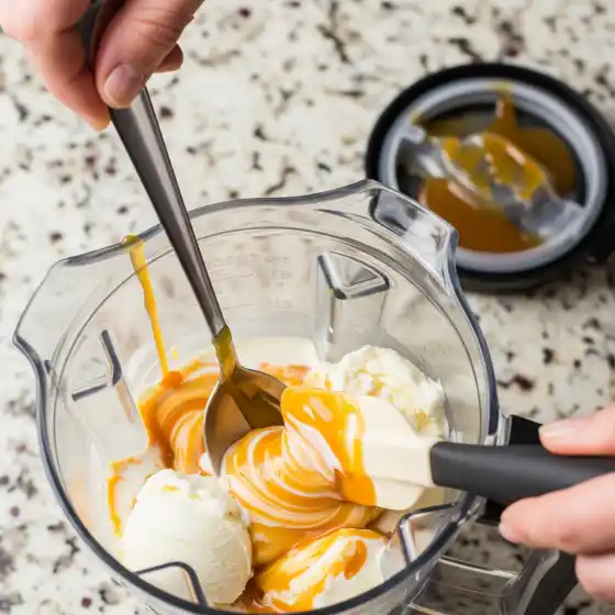 Two hands stirring vanilla ice cream and caramel sauce in a blender to prepare a Caramel Milkshake, with a lid in the background on a speckled countertop.