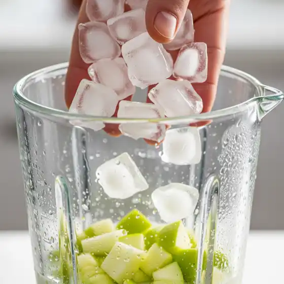 A hand adds ice cubes into a blender filled with green apple chunks, preparing an apple milkshake. The scene is bright and fresh, suggesting a smoothie preparation.