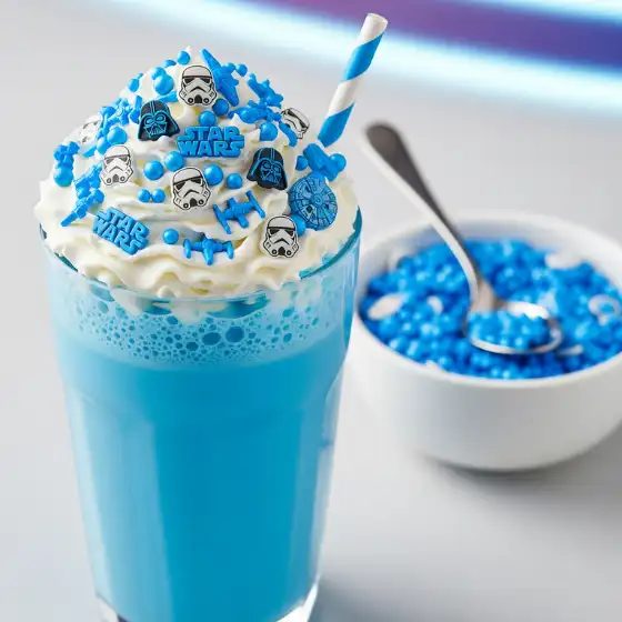A vibrant Blue Milkshake topped with whipped cream and blue Star Wars-themed decorations, including figures and logos. A matching bowl of decorations is nearby.