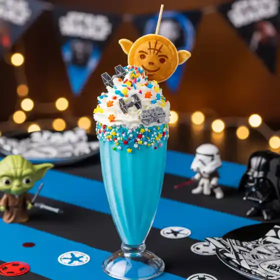 Blue Milkshake topped with whipped cream, colorful star sprinkles, and a cookie in the shape of a green character. Background shows themed banners and lights.
