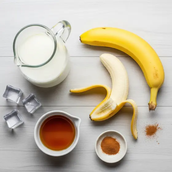 A flat lay of ingredients for a Banana Milkshake Recipe, featuring a pitcher of milk, two ripe bananas, honey, cinnamon, and ice cubes on a light wooden surface.
