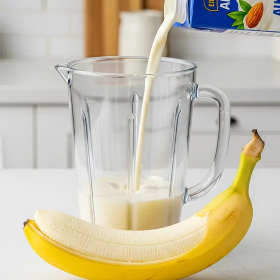 A banana lies beside a clear blender as almond milk is poured into it from a carton, ready for Peanut Butter Protein Shake preparation.
