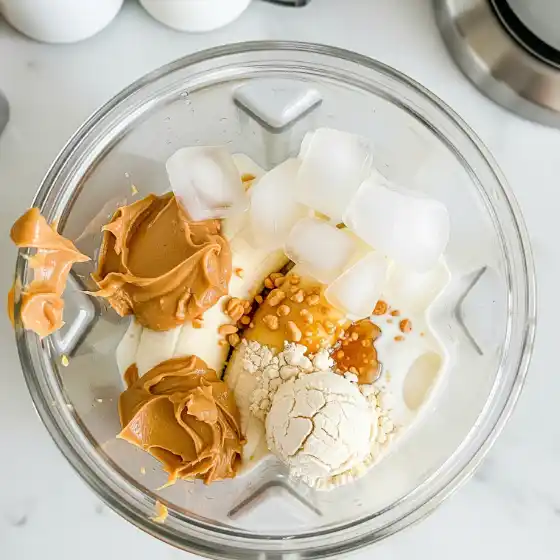 A blender filled with ice cubes, peanut butter, vanilla ice cream, and creamy ingredients for a delicious Peanut Butter Protein Shake smoothie.