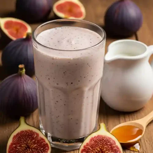 A creamy Fig milkshake in a tall glass on a wooden table, surrounded by fresh figs, a small white jug, and a spoonful of honey.